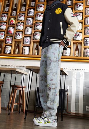 Osoba ubrana w czarno-beżową kurtkę varsity, jasne jeansy z nadrukiem i białe trampki, stojąca w pokoju z drewnianymi półkami trzymającymi ceramiczne słoje.