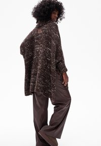 Kobieta z kręconymi włosami, nosząca brązowy sweter oversize z cekinami, szerokie brązowe skórzane spodnie i brązowe zamszowe buty, stojąca w pozycji bocznej.