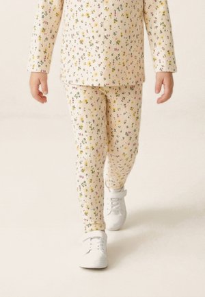 Gyerek krémszínű bordás hosszú ujjú felsőt és kis sárga, rózsaszín és zöld virágmintás nadrágot visel, fehér tornacipőben sétál.