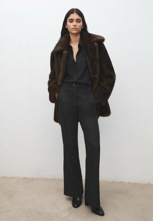 Brązowy futrzany płaszcz z ekologicznego futra na czarnej koszuli na guziki, zestawiony z czarnymi spodniami o wysokim stanem i czarnymi botkami na obcasie. Gładkie tekstury, dopasowany krój.