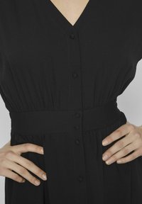 Čierne šaty s výstrihom do V, krátkymi rukávmi a sťahovacím pásom. Obsahuje zapínanie na gombíky a plisovanú sukňu. Hladká, ľahká látka.