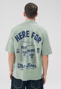 Light green button-up shirt with short sleeves featuring a large printed graphic of beer cans and text on the back.