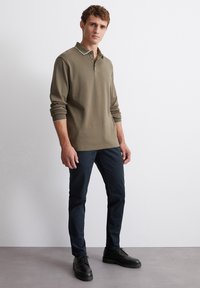 Olivgrünes Langarm-Polo mit Kragen und drei Knöpfen; kontrastierender weißer Besatz am Kragen und an den Ärmelbündchen; kombiniert mit dunklen Hosen und schwarzen Schuhen.