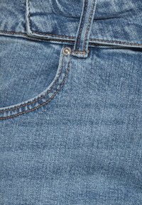 Primer plano de un bolsillo de jeans de mezclilla azul, mostrando el lazo del cinturón, el remache de metal y los detalles de costura naranja.