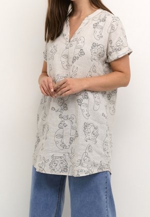 Woman wearing a beige tunic with abstract black line art faces and floral patterns, paired with blue denim jeans, standing against a light background.