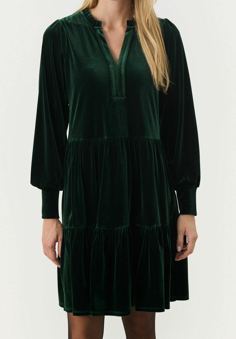 Femme portant une robe en velours vert foncé, à manches longues, à volants, longueur genou, avec une encolure fendue, sur un fond uni.
