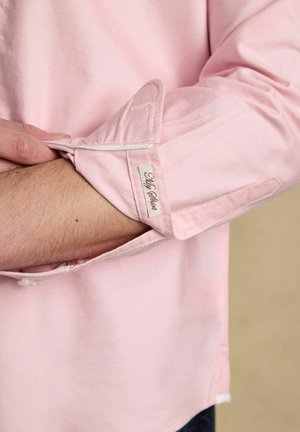 Person, die die Manschette eines hochgekrempelten, hellrosa Langarmhemds mit einem aufgenähten "My Shirt"-Etikett anpasst.