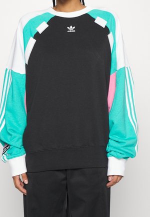 Persona che indossa una felpa nera con blocchi di colore teal, rosa e bianco, logo Adidas sul petto e tre strisce bianche sulle maniche teal.