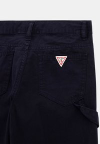 Marineblå bukser med en trekantet rød og hvid "GUESS"-logo på baglommen, med et klassisk design og en glat tekstur.