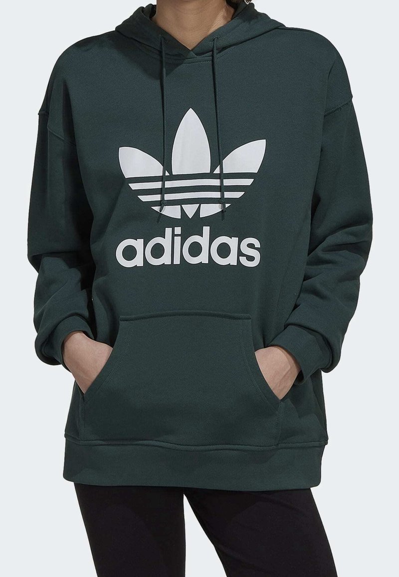 Grön hoodie med en känguruficka och justerbara dragskor. Har en stor vit Adidas-logotyp och ett mönster med tre ränder. Tillverkad av mjukt material.