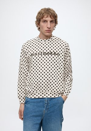 Ung person med krøllet hår iført hvid langærmet skjorte med sorte prikker og teksten ""marimekko"" samt blå jeans, står mod hvid baggrund.
