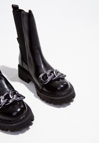 Bottines en cuir noir avec une semelle épaisse, présentant un accent en chaîne métallique sur le devant. Texture lisse et panneau latéral élastique.