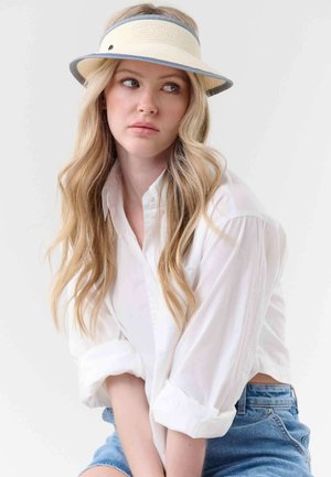 Młoda kobieta o długich blond włosach, ubrana w białą koszulę z guzikami, niebieskie szorty dżinsowe i słomkową daszek, patrząca w bok.