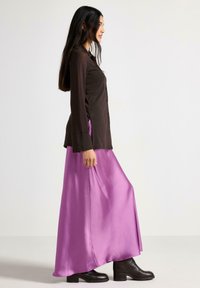 Kvinde i profil iført en sort langærmet skjorte, flydende skinnende lilla nederdel og sorte ankelstøvler mod en ensfarvet lys baggrund.