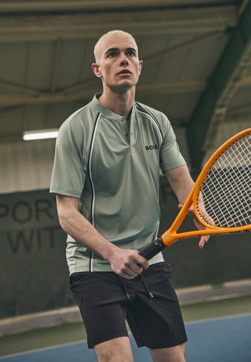 Jasnozielona koszulka polo z czarnymi akcentami, noszona z czarnymi szortami. Osoba trzyma pomarańczową rakietę tenisową, stojąc na korcie.