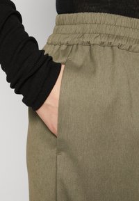 Olivgröna byxor med elastisk midja och sidoficka. Tyget har en slät textur och en lätt glans. Svart långärmad topp syns.