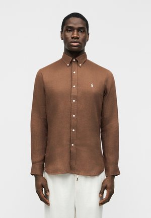 Mężczyzna ubrany w brązową, zapinaną na guziki koszulę z długim rękawem i kołnierzykiem, ozdobioną białym haftowanym logo, w połączeniu z białymi spodniami na jednolitym tle.