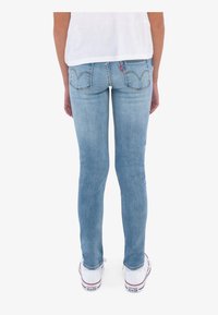 Persona que lleva jeans ajustados de color azul claro y zapatillas Converse blancas, vista desde atrás sobre un fondo blanco liso.