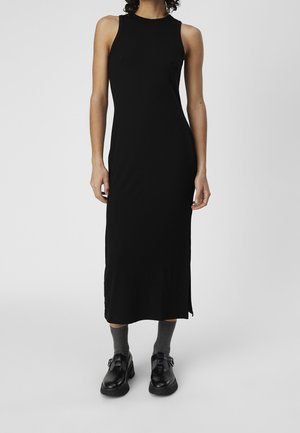 Frau trägt ein ärmelloses schwarzes Midikleid, graue Socken und schwarze Schuhe mit Schnallen, steht vor einfachem hellem Hintergrund.