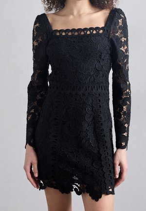 Femme portant une robe en dentelle noire à manches longues et un décolleté carré, avec des motifs floraux et tourbillonnants.