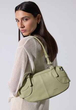 Mujer con cabello largo y oscuro viste un top de punto color beige claro y lleva un bolso de cuero verde pálido colgado del hombro.