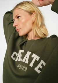 Olivgrön sweatshirt med rund halsringning och ribbade ärmslut, med "CREATE 2010" i stora, texturerade vita bokstäver på framsidan.