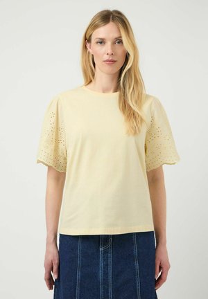 Blonde vrouw die een lichtgele blouse met opengewerkte pofmouwen draagt en een donkerblauwe spijkerrok, staand tegen een effen witte achtergrond.