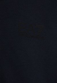 Tessuto a maglia blu navy con una trama liscia, caratterizzato da un logo in rilievo nero "EA7 Emporio Armani". Design semplice e pulito.