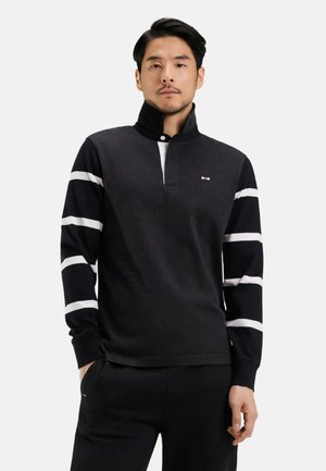 Homme portant un haut à manches longues noir à fermeture éclair quart avec des rayures blanches horizontalement sur les manches et un pantalon noir assorti, se tenant avec une main dans la poche.