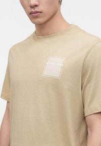 Νέος άνδρας που φοράει μπεζ κοντομάνικο T-shirt με μικρό τετράγωνο σχέδιο και το κείμενο "MICHAEL KORS" στο στήθος.