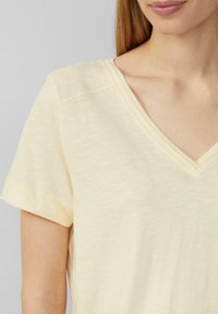 Jasnożółta koszulka z dekoltem w serek, krótki rękaw, lekko teksturowana tkanina, widoczne szwy, miękki spływ, luźny krój, minimalistyczny design.