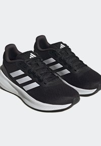 Zapatillas de running Adidas negras con suelas blancas y tres franjas blancas a los lados, presentadas lado a lado sobre un fondo claro.