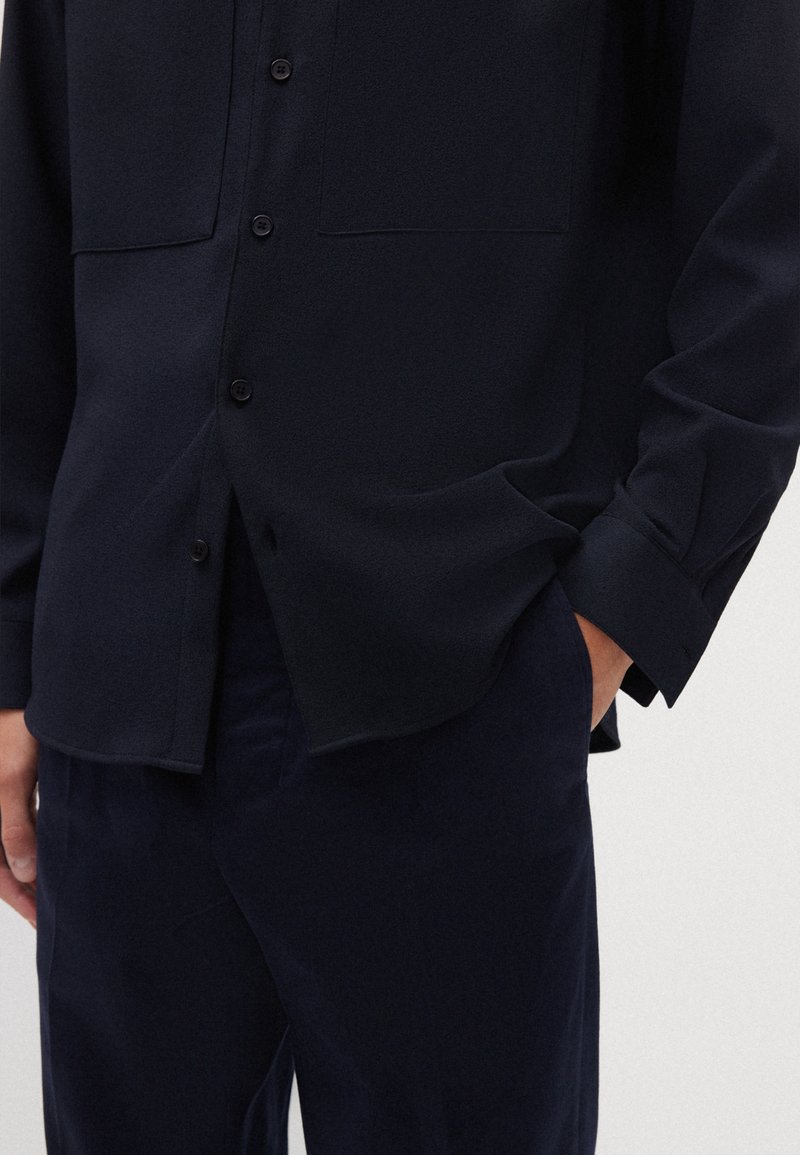 Navyblaues Button-Up-Hemd mit zwei Brusttaschen, langen Ärmeln und glatter Textur. Kombiniert mit passenden dunklen Hosen, die ein einfaches Design zeigen.