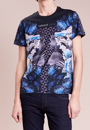 Man draagt een Versace Jeans T-shirt met een blauw bloemen- en geometrisch patroon, gecombineerd met een donkere spijkerbroek tegen een neutrale achtergrond.