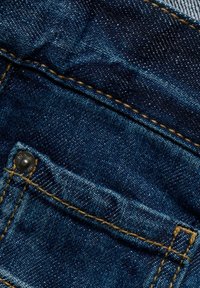 Mörkblå denimtyg med en ficka med en metallnitar. Kontrastsgarnering i gult, slät textur och lätt slitage synligt.