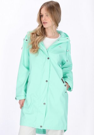 Nő, aki hosszú, kapucnis mentazöld esőkabátot visel, egyik kezét a zsebében tartva, kissé balra néz.