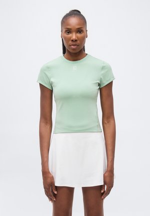 Sötét bőrű, hátra kötött hajú nő világoszöld, testhez simuló rövid ujjú felsőt és fehér szoknyát visel, egyszínű háttér előtt áll.