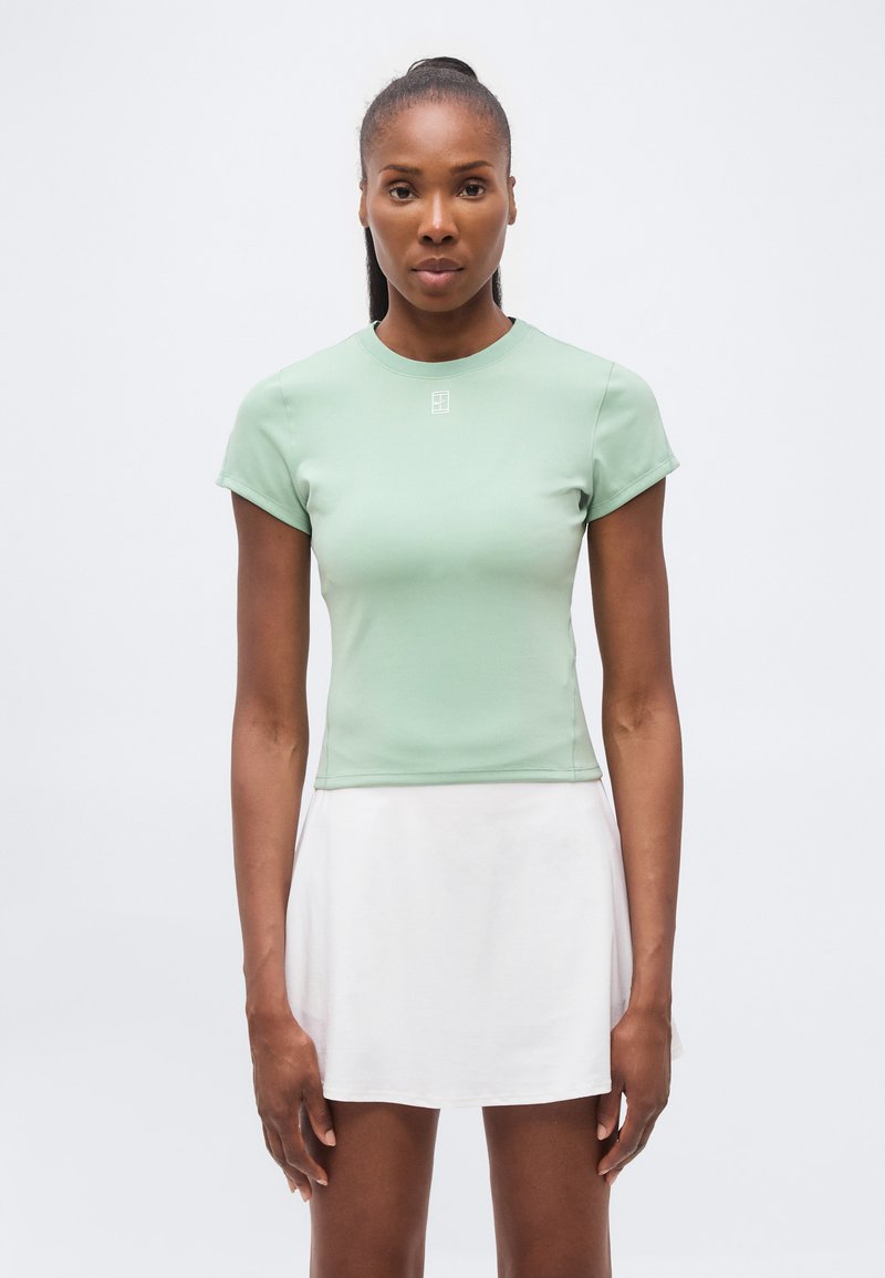 Γυναίκα με σκουρόχρωμο δέρμα και μαλλιά δεμένα πίσω, φορώντας ένα ανοιχτό πράσινο εφαρμοστό κοντομάνικο τοπ και λευκή φούστα, στέκεται μπροστά σε ένα μονόχρωμο φόντο.