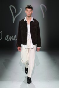 Tumši brūns zamšs jackets virs baltas krekla, apvienots ar krēmkrāsas biksēm un melniem apaviem. Models stāv pret maigi apgaismotu fonu.