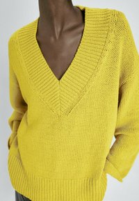 Pull jaune en maille à col en V avec col, poignets et ourlet côtelés, porté par une personne les mains dans les poches, sur fond uni.