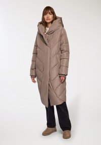 Kurtka puchowa w jasnym taupe z przeszyciami w zygzak, oversize'owy kaptur, długie rękawy i boczne kieszenie, w połączeniu z ciemnymi spodniami i beżowymi butami.