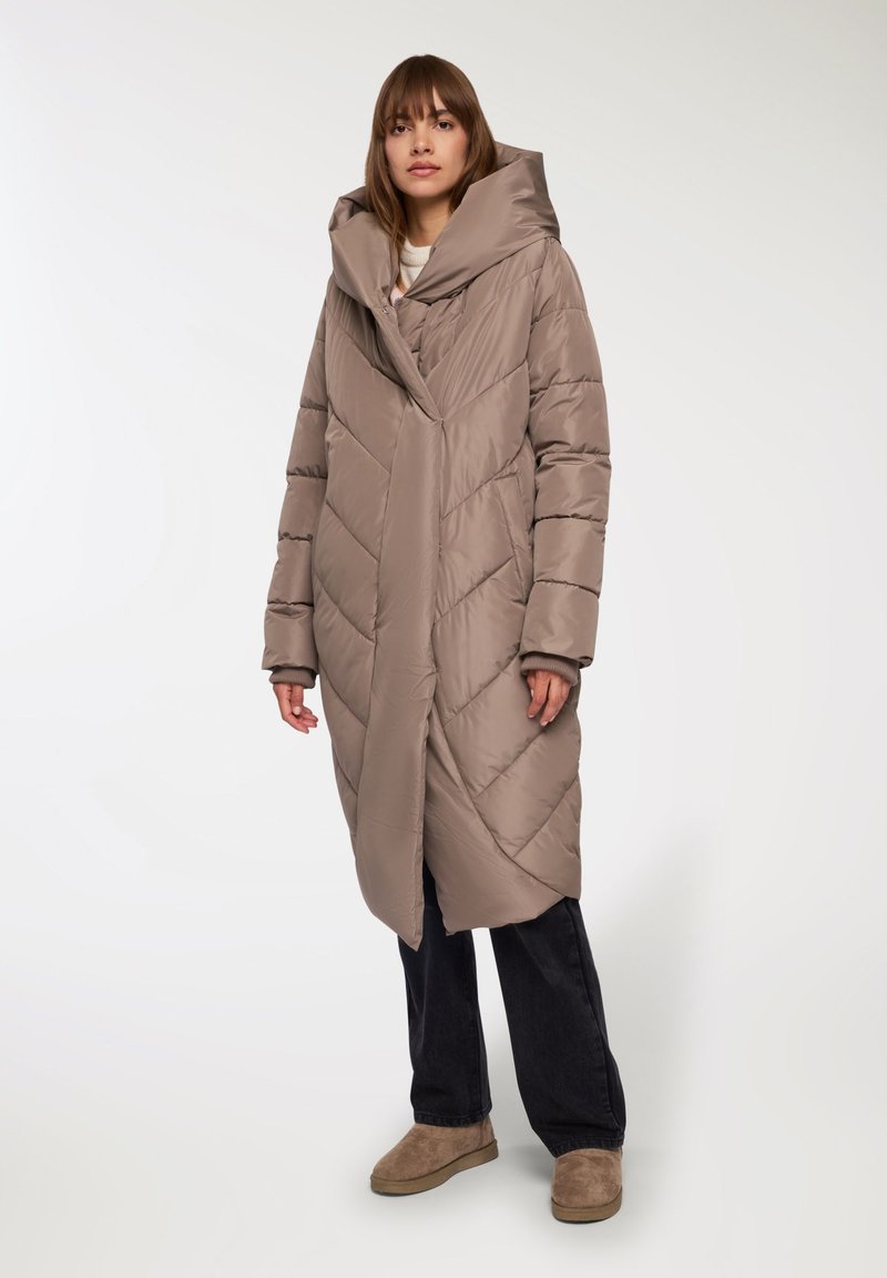 Kurtka puchowa w jasnym taupe z przeszyciami w zygzak, oversize'owy kaptur, długie rękawy i boczne kieszenie, w połączeniu z ciemnymi spodniami i beżowymi butami.