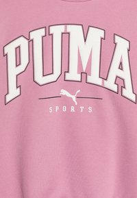 Sudadera rosa con gran texto blanco "PUMA" y "SPORTS" en letras más pequeñas debajo, con una silueta del logotipo de un puma. Textura de tela suave.