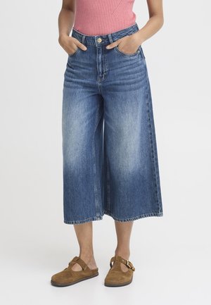 Person iført korte viddeblå jeans, lyserød ærmeløs top og brune slip-on sko med spænder, hænderne i lommerne.