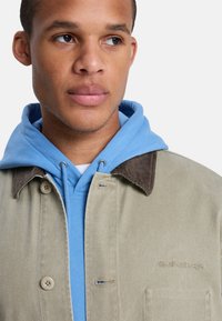 Ανοιχτό μπεζ μπουφάν με καφέ γιακά από κορντούρα, φορεμένο πάνω από μπλε φούτερ με κουκούλα. Διαθέτει κουμπιά στο εμπρός μέρος και κεντημένο λογότυπο στο μπουφάν.