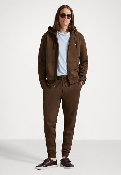 Sudadera marrón y joggers a juego hechos de tela suave, combinados con una camiseta de color azul claro. Zapatillas marrones con suelas blancas completan el atuendo.