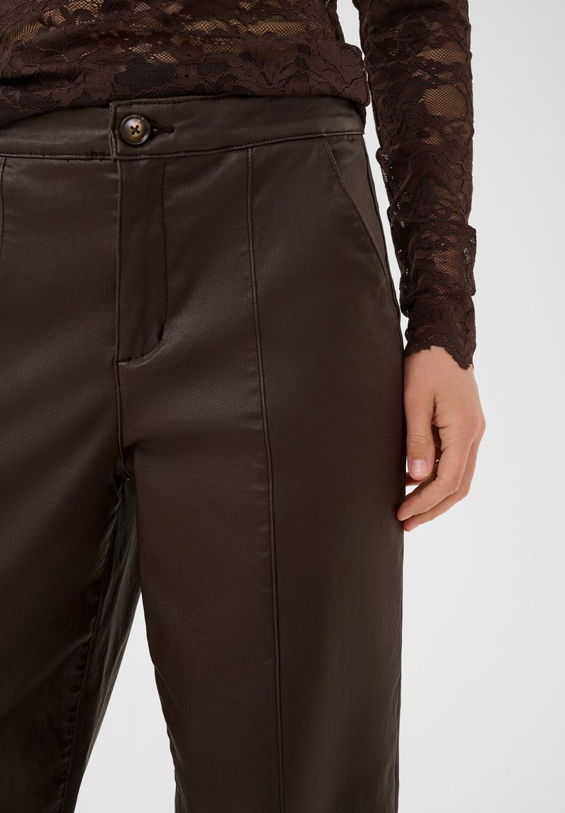 Personne portant un pantalon marron foncé brillant avec des poches latérales et un haut en dentelle marron à manches longues, la main droite reposant sur le côté.
