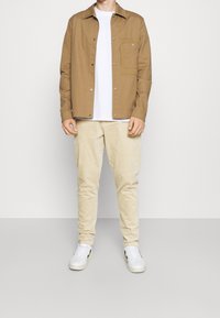 Chemise marron à boutons avec une poche poitrine, t-shirt blanc en dessous, pantalon crème et baskets blanches avec des accents noirs.