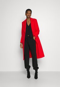 Roter langer Mantel mit taillierter Passform, kombiniert mit einem schwarzen Croptop und schwarzen Jogginghosen. Schwarze Stiefeletten runden das Outfit ab.