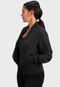 Schwarze Bomberjacke mit Reißverschluss vorne, gerippten Bündchen und seitlichen Taschen. Verfügt über einen strukturierten Patch auf der Brust und ein glattes, leichtes Gewebe.